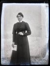 Portrait d'une femme en robe sombre debout, tenant le journal "Le petit journal" plié dans sa main.