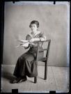 Portrait d'une jeune femme assise, accoudée au dossier de la chaise, une publication ouverte entre les mains.
