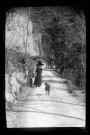 Femme se promenant sur un chemin au pied d'une paroi rocheuse, un chien l'attend.