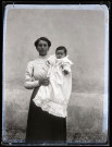 Portrait d'une femme debout tenant un bébé en tenue de baptême dans les bras.