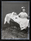 Portrait d'un bébé en tenue de baptême dans un landau à côté d'une petite fille.