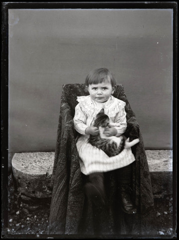 Portrait d'un petit enfant en robe rayée assis sur une chaise, un chaton dans les bras.