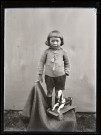 Portrait d'un garçon debout, avec à ses pieds un petit cheval de bois sur roulettes.