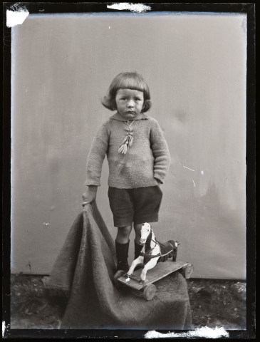 Portrait d'un garçon debout, avec à ses pieds un petit cheval de bois sur roulettes.