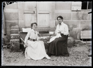 Portrait de deux femmes assises autour d'une table, affairées l'une à la lecture, l'autre à la couture, devant les marches du pensionnat de jeunes filles rue du Tatet à Vers-en-Montagne.