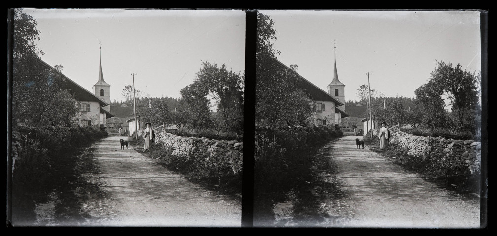 Rue de village, une dame et un chien posent près d'un muret de pierres.