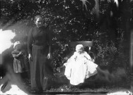 Une femme avec deux enfants en extérieur, dont l'un posé sur une chaise en habit de baptême