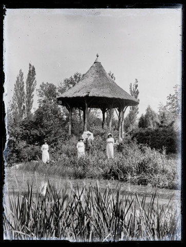 Trois femmes en blanc et un homme tenant une ombrelle devant un kiosque proche d'un plan d'eau.