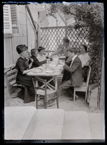 Six personnes déjeunant attablées dans le jardin d'une maison. Anne Coutemoine est assise à gauche, en face de ses enfants Jean Rameaux et Agathe Rameaux.