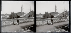Femme sur un chemin le long d'un muret en pierre, une église et des bâtiments en arrière-plan.