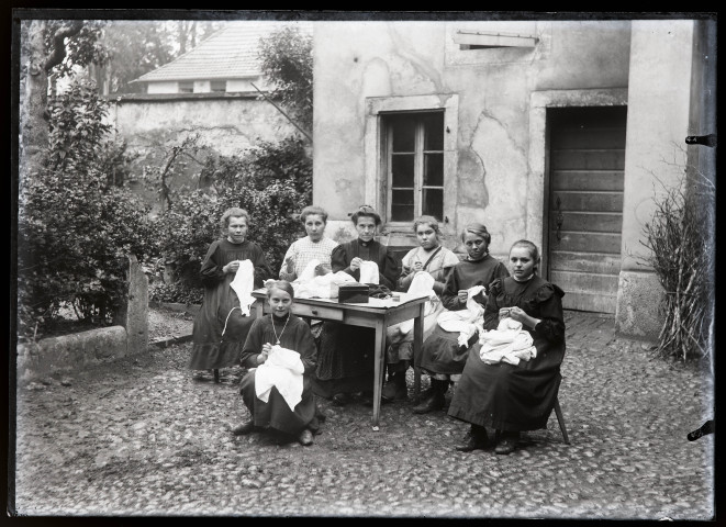 Femme apprenant à coudre à un groupe de jeunes filles dans la cour de l'école Saint-Laurent de Vers-en-Montagne.