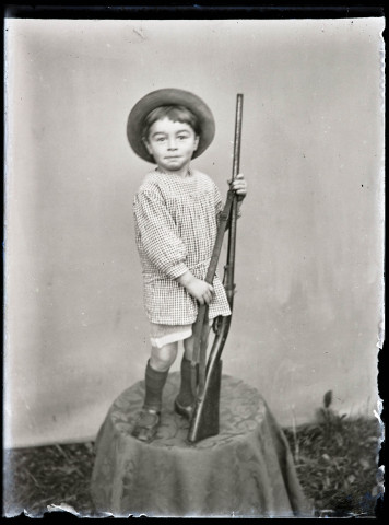 Portrait d'un petit garçon debout, avec chapeau et carabine.