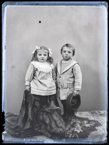Portrait d'une petite fille assise à côté d'un petit garçon un chapeau à la main, les deux enfants se tiennent par la main.