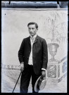 Portrait de Jean Rameaux tenant canne, gants et canotier, posant devant un décor peint.