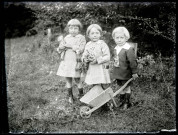 Portrait en extérieur d'un petit garçon poussant une brouette accompagné de deux fillettes tenant l'une une poupée et l'autre un bouquet de fleurs.