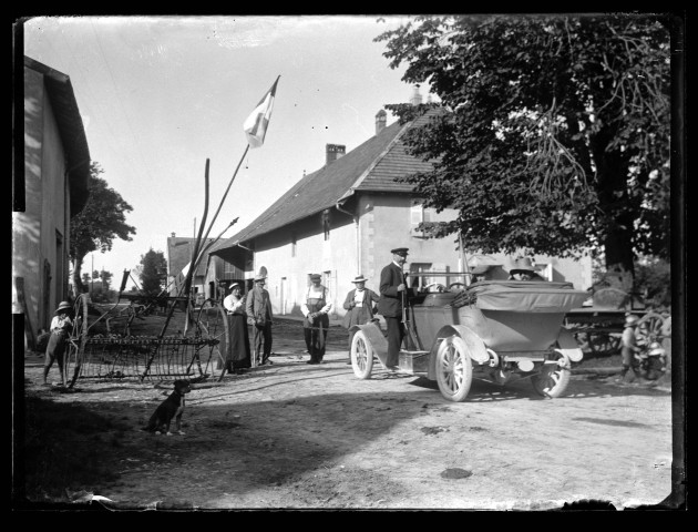 Hommes et femme armés autour d'une voiture semblable à la célèbre "voiture Pichon" dans une rue pavoisée de Vers-en-Montagne.