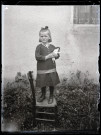 Portrait d'une petite fille tenant une poupée dans les bras, debout sur une chaise en extérieur.