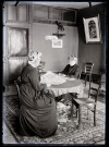 Deux femmes assises autour d'une table dans une habitation, l'une lit le journal "L'écho de Paris", l'autre écrit.