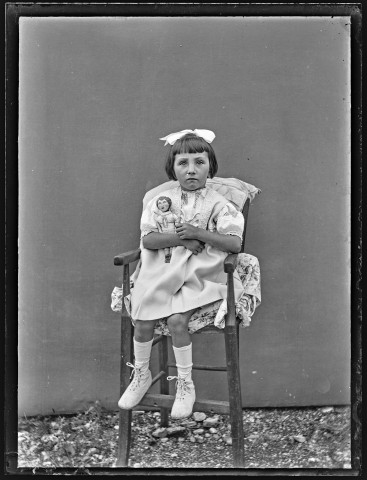 Portrait d'une petite fille assise sur une chaise, une poupée dans les bras.