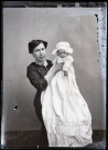 Portrait d'une femme assise tenant dans les bras un bébé en tenue de baptême.