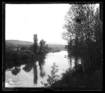 Paysage du Doubs avec un cours d'eau, des arbres, des maisons et un pont au loin.
