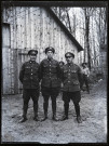 Portraits du Corps des forestiers canadiens et autres troupes : trois militaires en uniforme posant devant un baraquement en bois, deux bucherons en arrière-plan.