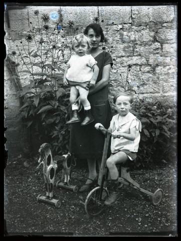 Portrait de femme portant un petit enfant, un jeune garçon sur un tricycle et un cheval de bois à ses pieds.,